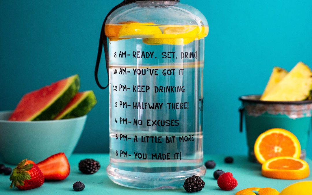 Photo of a clear water with markings on the side to delineate times of the day to drink water for optimal hydration, the bottle filled with water and citrus slices